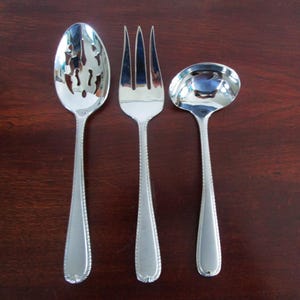 May include: A set of three silver-colored serving utensils: a slotted spoon, a fork, and a ladle. Each piece has a decorative beaded edge and a polished, reflective surface. The utensils are arranged on a dark brown wooden surface.
