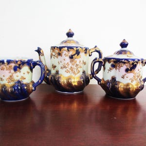 Może przedstawiać: Zestaw do herbaty z porcelany vintage z złotymi akcentami. Czajnik, dzbanek na śmietanę i cukiernica są ozdobione kwiatowymi wzorami i mają niebiesko-białą podstawę.