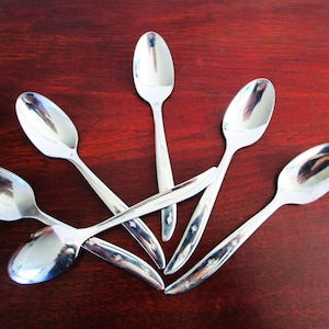 May include: Five silver spoons arranged in a fan shape on a dark red wooden surface. The spoons have a simple design with a rounded bowl and a slightly curved handle.