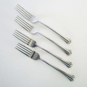 May include: A set of four silver-toned metal forks arranged diagonally. Each fork features four tines and a handle with a flared, scalloped base design. The polished finish reflects light, highlighting the smooth curves of the metal construction.