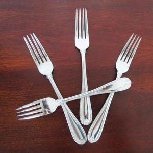 May include: Five silver forks arranged on a dark wood surface. The forks have a classic design with tines and ornate handles. The arrangement creates a star-like pattern, showcasing the silverware.