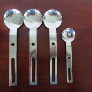 May include: Four stainless steel spoons with a distinctive design. The spoons have a circular bowl and a long, flat handle with a hole and a rectangular cutout. The spoons are arranged on a dark brown surface.