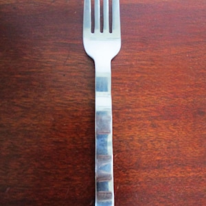 May include: A stainless steel fork with four prongs. The handle features a textured, rectangular pattern. The fork is placed on a dark brown wooden surface, providing a contrast in colour and texture.