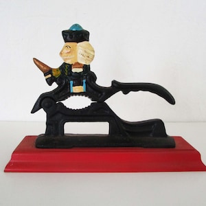 Op de afbeelding: Vintage notenkraker van gietijzer met een zwart hefboommechanisme, gemonteerd op een rode rechthoekige basis. De geschilderde figuur draagt een hoed en een jas. De figuur houdt een klein voorwerp vast.