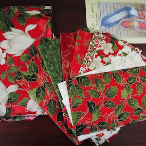 Op de afbeelding: Een verzameling kerststoffen in verschillende patronen. De stoffen hebben rode achtergronden met hulst, bessen en bloemmotieven. Een patroonblad voor een tafelloper is ook inbegrepen, met de tekst "Braid Runner".
