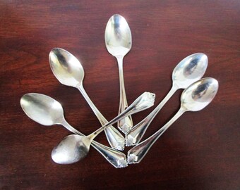 以下が含まれることがあります： 濃い茶色の木製の表面に、扇形に並んだ銀製のティースプーン5本