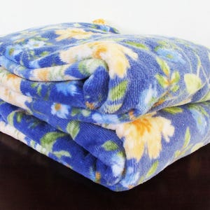 Puede incluir: Una pila de tres mantas azules plegadas con un estampado floral. El estampado presenta flores amarillas y blancas con hojas verdes. Las mantas están apiladas sobre una superficie marrón oscuro.