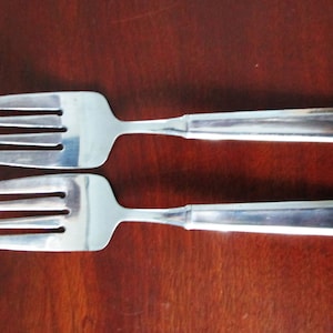 May include: Two silver forks with simple, squared handles. The forks are laid out parallel to each other on a wooden surface.