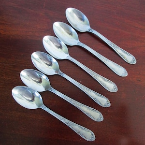 May include: Six silver spoons arranged diagonally on a dark wood surface. The spoons have oval bowls and ornate, engraved handles. The spoons are reflective, showing the surrounding environment.
