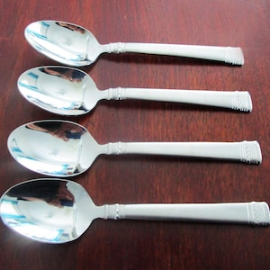 May include: Four silver spoons with oval bowls and detailed handles are arranged on a dark wooden surface. The spoons have a reflective, polished finish, and the handles feature a decorative design near the bowl and at the end.