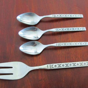 Op de afbeelding: Vier zilveren lepels en een zilveren vork met een decoratief patroon op de handvatten. De lepels hebben een ronde kom en de vork heeft drie tanden.