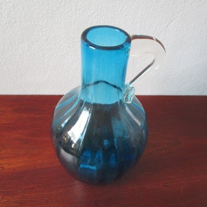 May include: A blue glass vase with a clear glass handle. The vase has a rounded body and a textured surface.