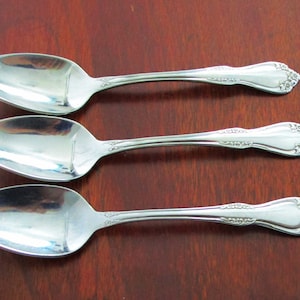 May include: Three silver spoons with ornate handles on a wooden surface.