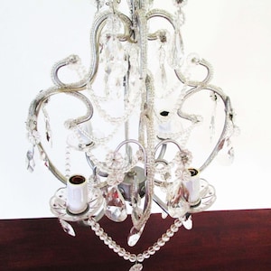 May include: A silver-toned metal chandelier featuring curved arms and multiple white candle-style light sockets. The fixture is adorned with clear glass crystal droplets and strings of small clear beads hanging from the frame in a draped pattern.