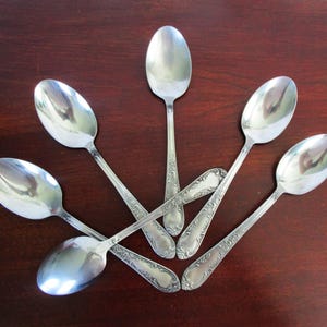 May include: Six silver spoons arranged in a starburst pattern on a dark wood surface. The spoons have a reflective, polished finish and ornate floral detailing on the handles. The spoons are a classic silverware design.
