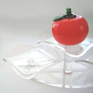 Puede incluir: Una bandeja transparente de varios niveles con una manzana roja de cristal encima. La manzana tiene un tallo y una hoja verdes. La bandeja es de acrílico transparente y tiene un diseño ondulado único. Ideal para exhibir objetos pequeños.