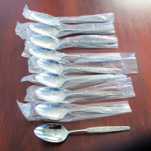 Puede incluir: Un juego de diez cucharaditas de plata con un diseño adornado. Las cucharas están envueltas individualmente en plástico transparente y están dispuestas en una fila sobre una superficie de madera.