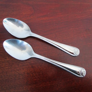 May include: Two shiny, silver spoons with a decorative design at the handle's end. The spoons are placed on a dark brown wooden surface. The spoons are likely for eating.