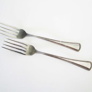May include: Two silver forks with a classic design are displayed against a white background. The forks have three tines and a handle with a beaded border. The handles have a rounded, teardrop shape.