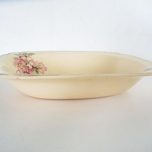 Puede incluir: Cuenco de cerámica ovalado de color crema con borde festoneado y diseño floral. El cuenco presenta un delicado patrón de flores rosas y hojas verdes. Un fino borde plateado perfila el borde.