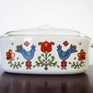 May include: A white oval casserole dish with a clear glass lid, featuring a colourful graphic print of two blue birds facing a central red tulip, surrounded by small red and orange flowers and green stems on the exterior of the white ceramic body.