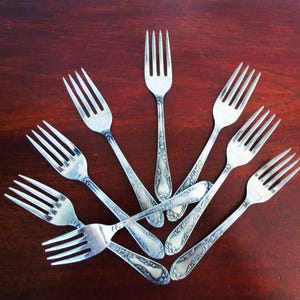 Puede incluir: Una colección de nueve tenedores plateados dispuestos en forma de abanico sobre una superficie de madera oscura. Los tenedores tienen un diseño clásico con púas y mangos ornamentados. La disposición crea una exhibición visualmente atractiva de cubiertos.