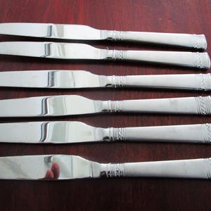 May include: Six stainless steel butter knives with serrated blades and decorative handles are arranged on a dark wood surface. The knives are reflective, catching the light, and appear to be part of a silverware set.