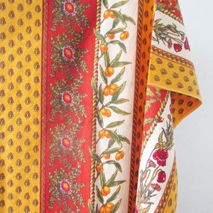 May include: A rectangular fabric textile featuring vertical panels with repeating patterns. The design includes yellow sections with small red motifs, a red band with floral elements, and a cream-colored stripe displaying orange fruit and green leaves.