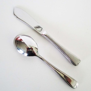 May include: A silver-toned metal butter knife and a matching spoon, both featuring a polished finish and a decorative beaded edge detail along the handle. The items are arranged diagonally, showcasing the reflective surface and the intricate trim.