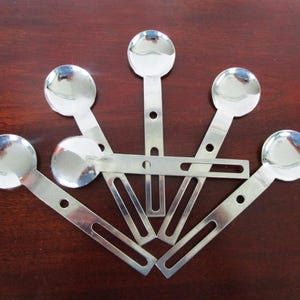 May include: Six shiny, silver metal spoons with a circular bowl and a flat, rectangular handle. Each handle has a small hole and a slot. The spoons are arranged in a fan shape on a dark brown surface.