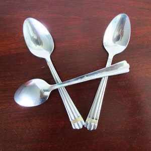 May include: Three silver spoons with gold accents are arranged in a triangular shape on a dark wood surface.