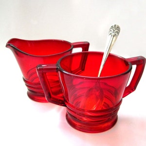 Può includere: Un set di brocca per la panna e zuccheriera in vetro rosso con un cucchiaio d'argento. La brocca per la panna e la zuccheriera hanno un design a coste.