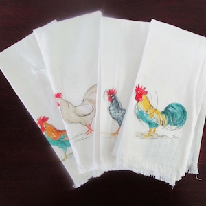 May include: A set of four white fabric napkins, each featuring a distinct illustration of a rooster. The roosters are depicted in various colours including red, yellow, blue, and grey. Each napkin has a fringed edge along the bottom hem.