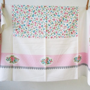 May include: A vintage white tablecloth with a floral pattern. The center features a dense arrangement of small pink and blue flowers. Pink borders with floral medallions and a black decorative trim frame the design. The tablecloth is hanging from a clothesline.