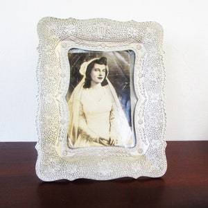 May include: A silver picture frame with a decorative floral design. The frame holds a black and white photograph of a woman in a wedding dress.