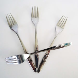 May include: Four silver forks arranged on a white surface. One fork is positioned horizontally across the others. The handles have a floral design. The forks are a kitchen utensil.