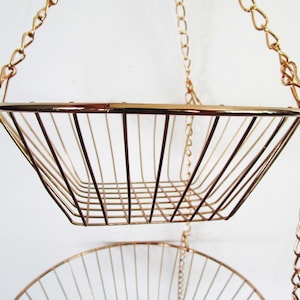 Puede incluir: Una cesta colgante de dos niveles hecha de alambre de metal en tono oro rosa. La estructura cuenta con dos cestas de marco abierto conectadas por cadenas de metal a juego, diseñadas para suspensión vertical con una base de rejilla.