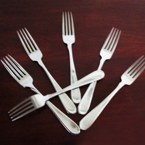 May include: Seven silver forks are displayed on a dark brown surface. Each fork features a three-tine design and a smooth, rounded handle. The forks are arranged in a radial pattern, creating a visually appealing arrangement.
