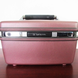 Pode incluir: Uma maleta de toucador rígida de cor malva vintage com acabamento texturizado, apresentando uma pega superior preta e um mecanismo de fecho central com detalhes cromados e a marca Samsonite. A mala retangular possui cantos arredondados.