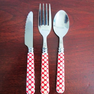 May include: A stainless steel knife, fork, and spoon set. The handles are red with white polka dots. The silverware is arranged on a dark brown surface.