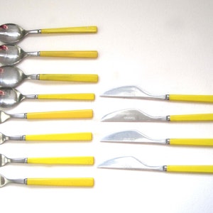 May include: A set of stainless steel flatware featuring bright yellow plastic handles. The collection includes four spoons, four forks, and four knives with curved blades, all arranged in a grid pattern against a plain white background.