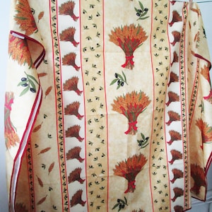May include: A beige tablecloth with a repeating pattern of wheat bundles tied with red ribbons, olive branches, and vertical stripes. The edges are trimmed with a red border. The tablecloth is hanging on a clothesline.