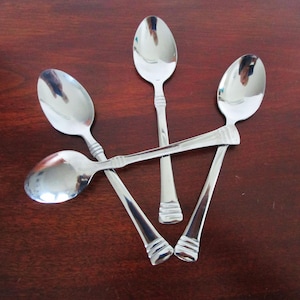 以下が含まれることがあります： 濃い茶色の木製の表面に配置された、光沢のあるステンレス鋼のスプーン5本。スプーンは楕円形のボウルと装飾的なハンドルが付いています。スプーンは光と周囲の環境を反射しています。