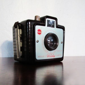 May include: Vintage Kodak Brownie Holiday camera. The camera is black with a light blue faceplate and red Kodak logo. The camera has a lens and a viewfinder. The camera is sitting on a dark brown surface.