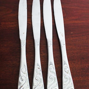 May include: Four silver butter knives with a decorative, scrolled design on the handles. The knives are arranged on a dark wooden surface, showcasing their polished blades and ornate details. Each knife is approximately 8 inches long.