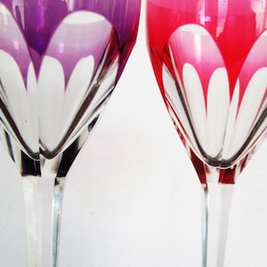 May include: Two elegant wine glasses with clear stems and bowls. One glass is a vibrant purple, the other a rich red. Both feature a white, petal-like design within the colored glass, creating a striking contrast.