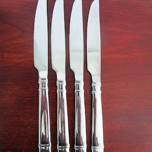 Può includere: Quattro coltelli da tavola in acciaio inossidabile con lame seghettate e manici ornati. I coltelli sono disposti verticalmente su una superficie di legno scuro, che riflette la luce. I manici hanno un design dettagliato e strutturato.