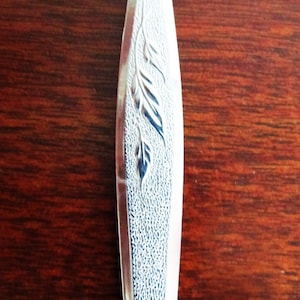 Può includere: Primo piano di un cucchiaio d'argento con un disegno a foglie. Il manico del cucchiaio ha una superficie strutturata e un motivo decorativo a foglie. Il cucchiaio è su una superficie di legno scuro.