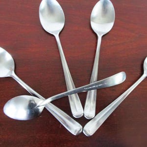May include: Five stainless steel spoons arranged on a dark brown wooden surface. The spoons have oval bowls and long, slender handles. The spoons are reflective, and the lighting creates highlights on their surfaces.