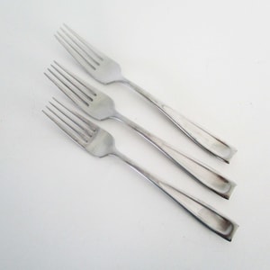 May include: Three silver-colored metal dinner forks arranged diagonally. Each fork features four tines and a smooth, slightly concave handle that tapers toward the end. The finish is polished and reflective, showing a clean, simple design.